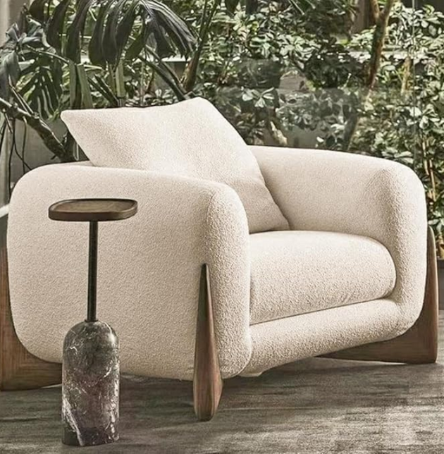 A modern living room featuring a large, plush, beige armchair, a dark marble side table, and green plants in the background.
