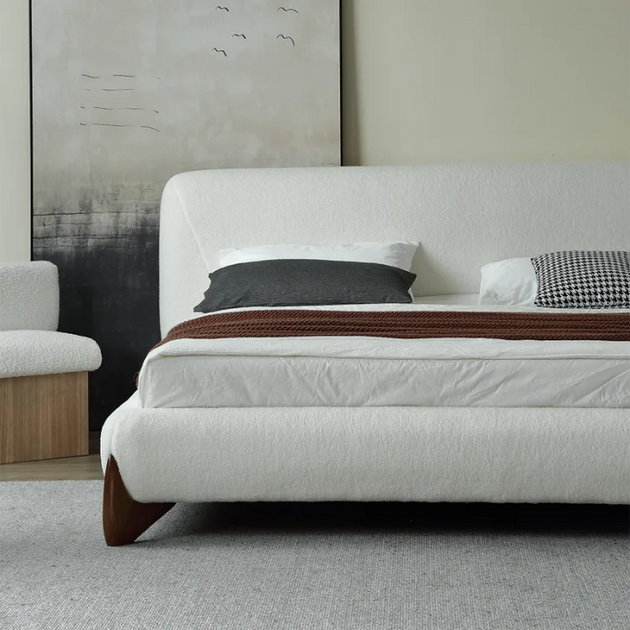 Contemporary low-profile bed frame upholstered in textured white or cream boucle fabric, featuring a rounded headboard and resting on a dark wood foot, set against abstract art.