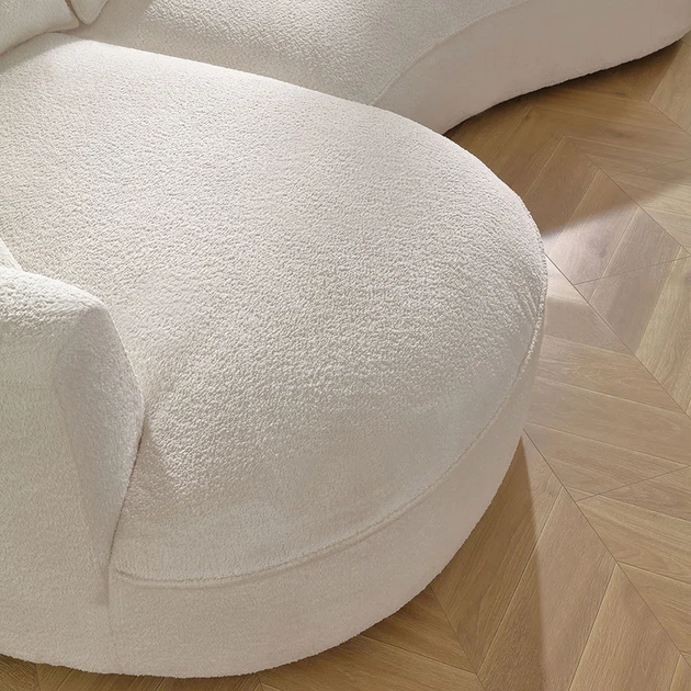Close-up detail of a curved, plush, white or cream-colored boucle fabric sofa or chair, resting on a light wood floor with a chevron or herringbone pattern