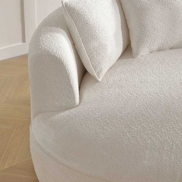 Close-up detail of a curved, plush, white or cream-colored boucle fabric sofa or chair, showing the texture and two loose back cushions.