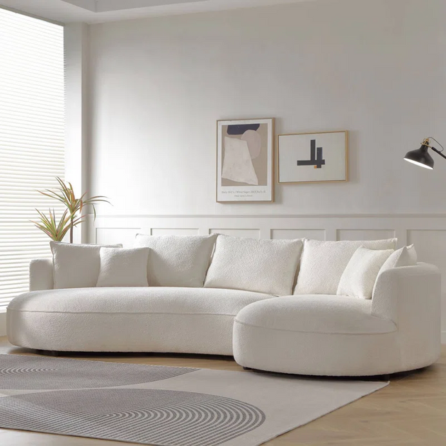 Curved, white boucle sectional sofa with five back cushions, placed on a light wood floor with a grey area rug in a modern living room featuring two framed abstract art prints.