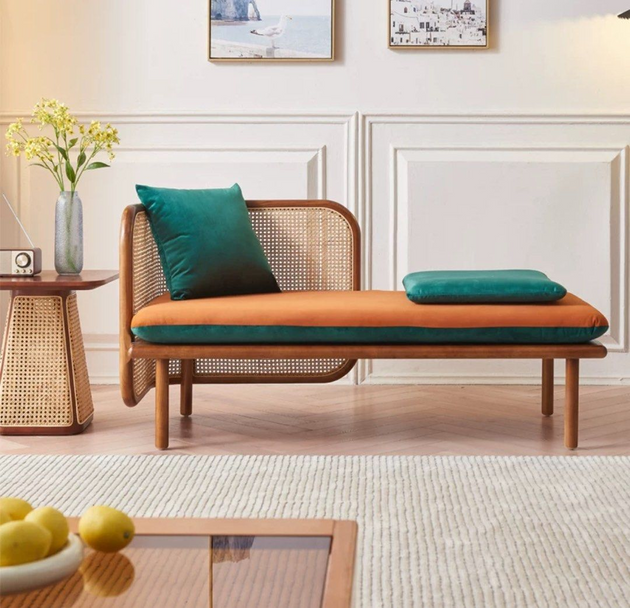 Modern rattan-frame daybed with orange cushioning and teal pillows, placed beside a woven side table with a vase of yellow flowers in a bright, minimalist living room.