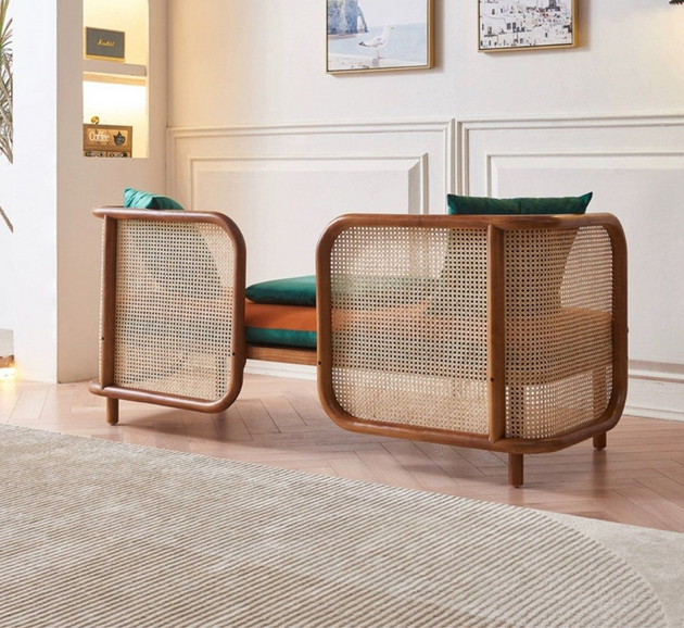 Side view of a modern rattan-frame sofa with orange cushions and teal pillows in a bright living room featuring white paneled walls and wooden flooring.